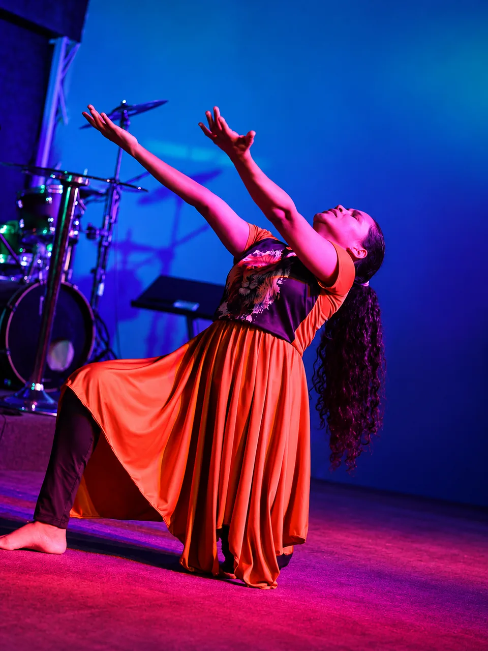 Dançarina de vestido laranja com os braços erguidos no palco, em adoração