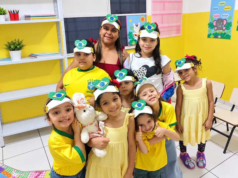 Crianças do MUP Kids de camiseta amarela e tiaras de coração verde e amarelo, sorrindo com a professora na sala