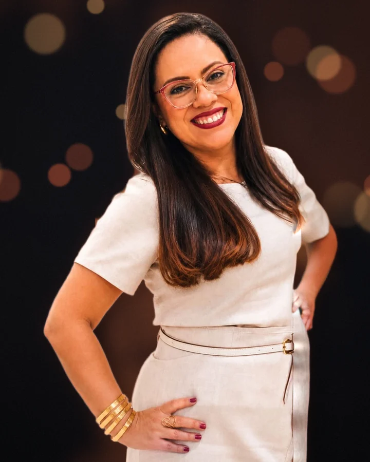Pastora Alessandra Mota sorrindo, de vestido claro e mãos na cintura, sobre fundo escuro de palco com luzes quentes