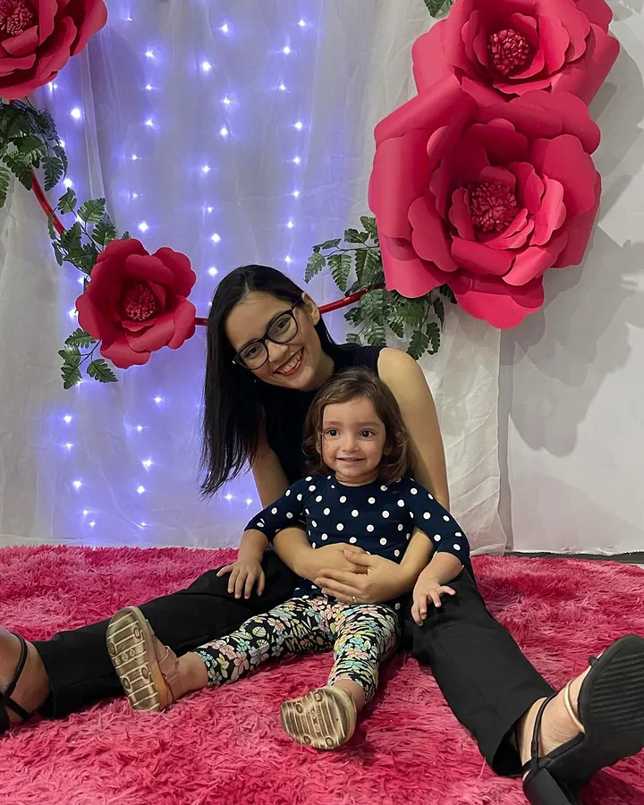 Mãe e filha pequena sentadas no tapete diante de um painel de rosas vermelhas